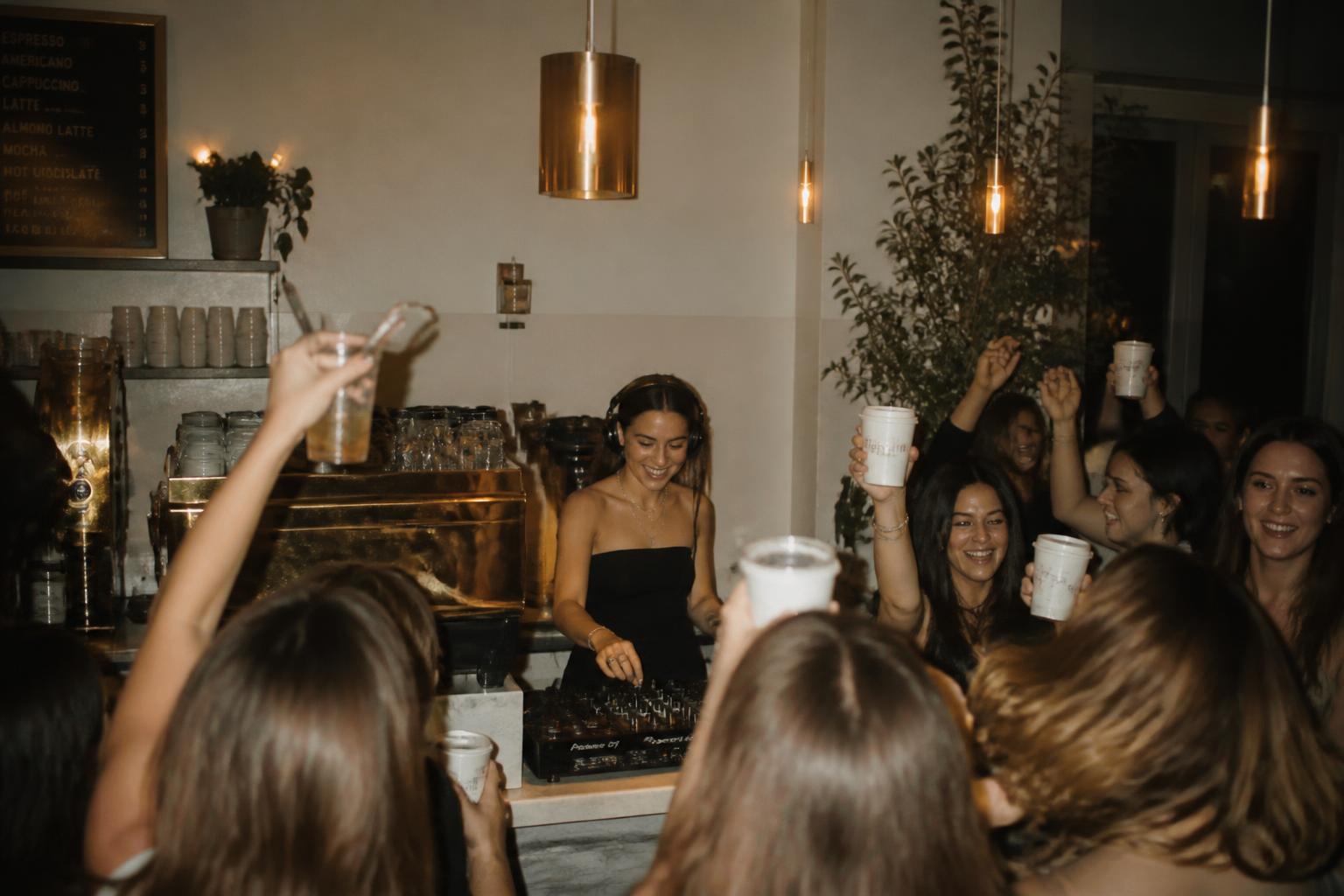 Candid photograph of a female DJ and a warm café crowd holding takeout coffee cups at a golden-lit coffee party